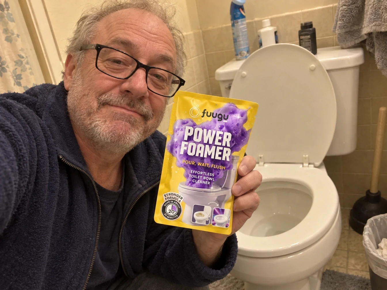 Phone selfie of a grey-haired man in glasses holding a Fuugu Power Foamer pouch beside his toilet in a beige-tiled bathroom